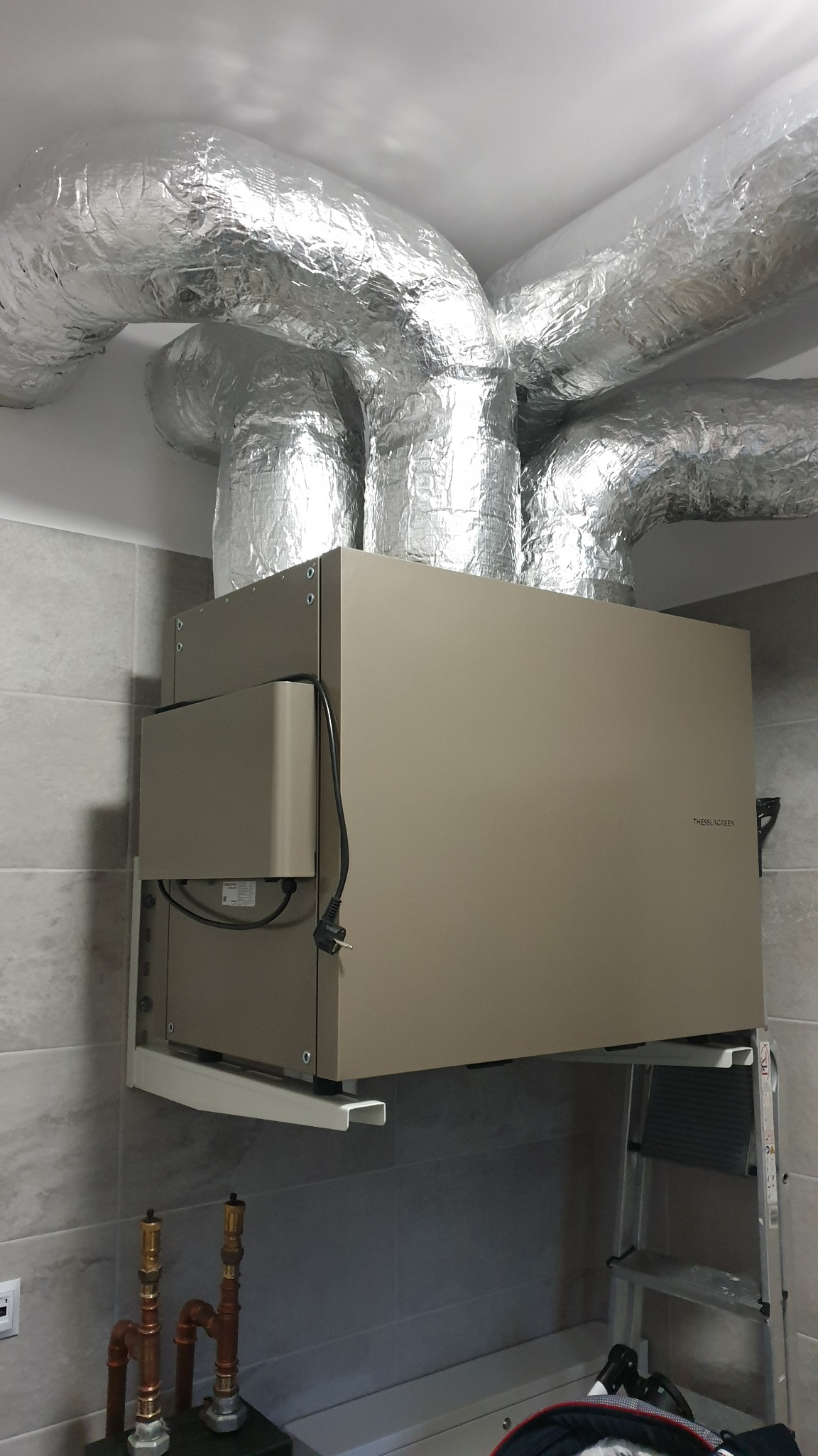 Szaro-beżowa jednostka rekuperacji Thessla Green zamontowana na ścianie z szarych płytek, z widocznymi izolowanymi przewodami wentylacyjnymi i fragmentem drabiny aluminiowej.