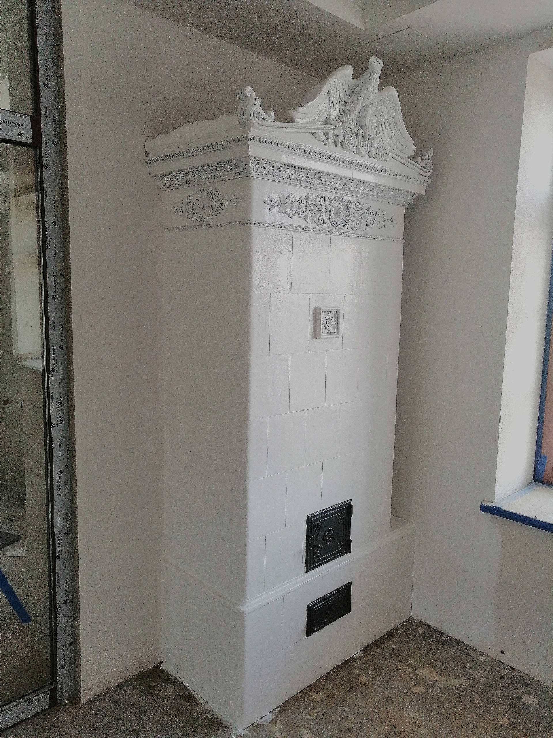 Biały, kaflowy piec z ozdobnym zwieńczeniem w postaci rzeźb orłów, umieszczony w surowym wnętrzu z widoczną stolarką okienną i betonową posadzką.