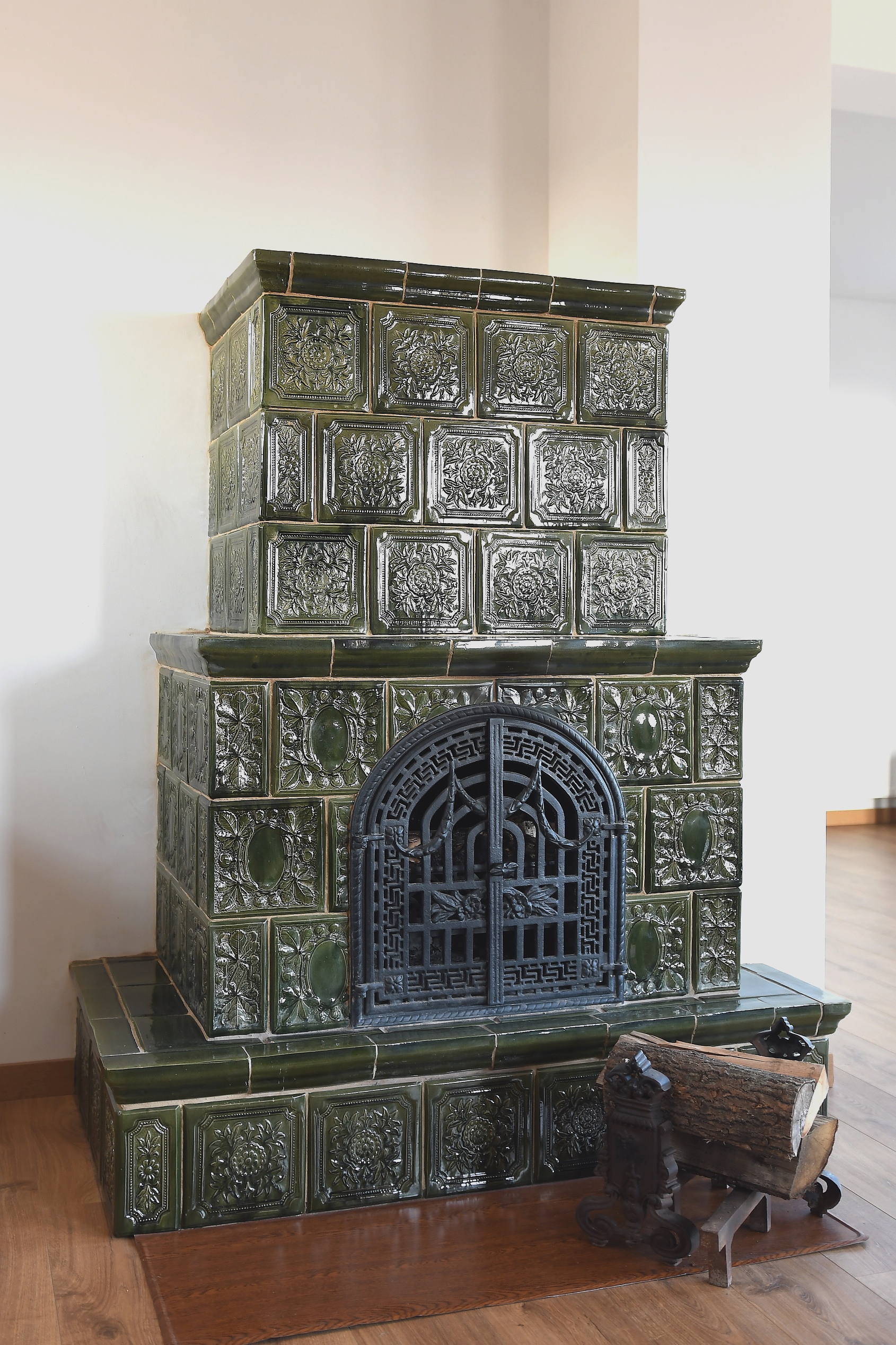 Zielony piec kaflowy z ozdobnymi kaflami i żeliwnymi drzwiczkami, obok leży drewno na ozdobnym stojaku.
