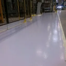 Jasnoszara, błyszcząca podłoga w hali magazynowej z metalowymi barierkami ochronnymi i wysokimi regałami w tle.