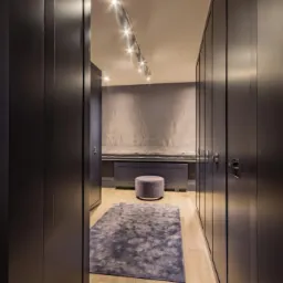 Garderoba w odcieniach szarości z oświetleniem punktowym, pufą i dywanem. Szafy z ciemnego drewna z metalowymi uchwytami po obu stronach korytarza.