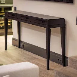 Elegancki, ciemny konsoletowy stolik z trzema szufladami, ustawiony przy ścianie z kontaktami. W tle widoczny regał na wino i fragment schodów.
