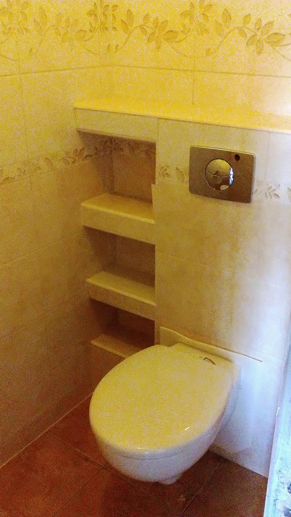 Beżowa toaleta podwieszana z półkami w ścianie, wykończona płytkami z motywem roślinnym. Widoczny przycisk spłuczki i brązowa podłoga.