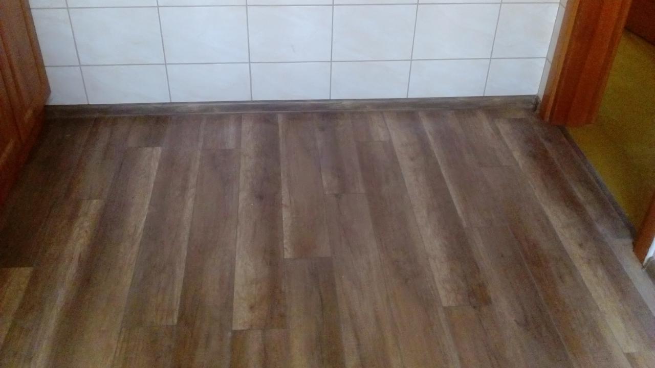 Ułożona, brązowa podłoga laminowana w pomieszczeniu z białymi płytkami na ścianie i drewnianymi listwami przypodłogowymi. Widok z góry, bliski kadr.