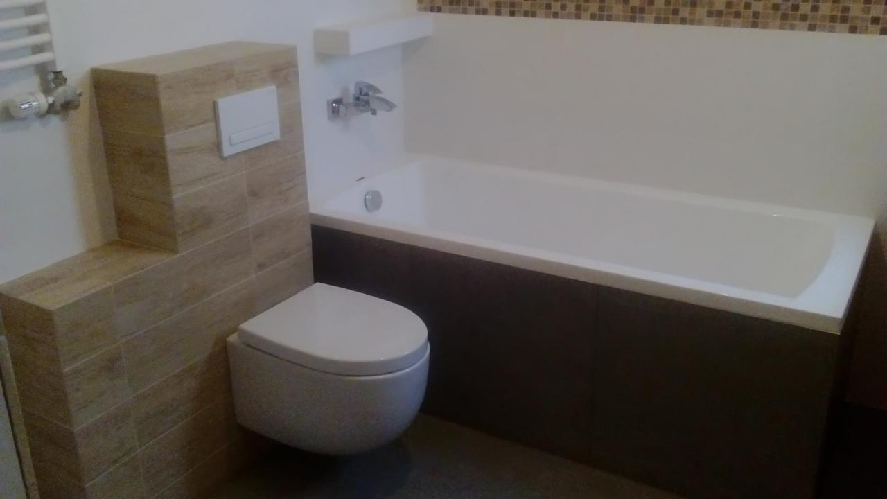 Nowoczesna łazienka z podwieszaną toaletą i wanną obudowaną ciemnymi płytkami. Uwagę zwraca mozaika nad wanną i płytki imitujące drewno.