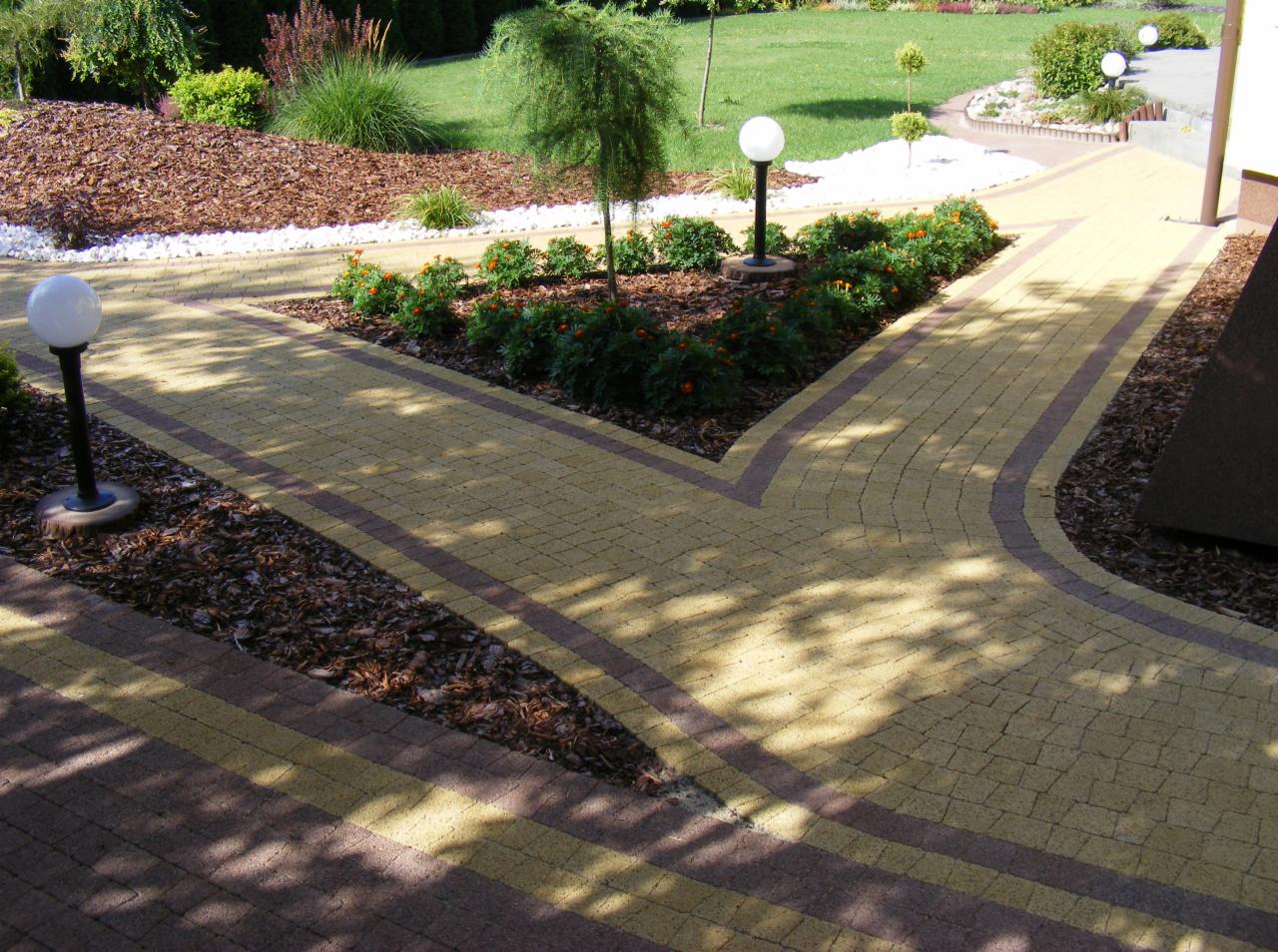 Żółto-brązowa kostka brukowa tworząca ścieżkę w ogrodzie, z elementami ozdobnymi: lampy, krzewy, kora. Widok na zadbany trawnik i elementy architektury ogrodowej.