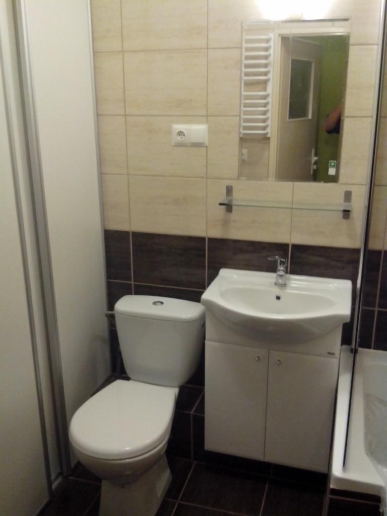 Wyremontowana łazienka w Bydgoszczy z białą toaletą, umywalką z szafką i lustrem, nad którym wisi grzejnik. Na ścianach beżowe i brązowe płytki.