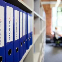 Rząd niebieskich segregatorów z etykietami 'TAXFORM.PL' na białych półkach biurowych, z widocznym w tle biurem i pracownikiem przy komputerze.