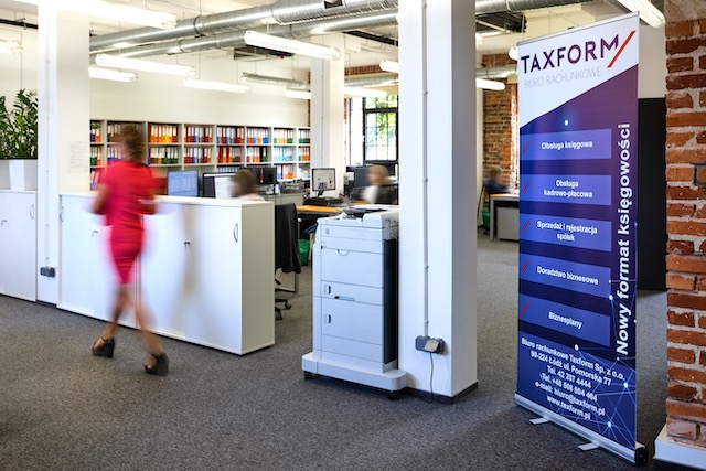 Wnętrze biura rachunkowego TAXFORM z regałami pełnymi segregatorów, drukarką i roll-upem reklamowym z ofertą usług, w tle widoczna pracownica w czerwonej sukience.