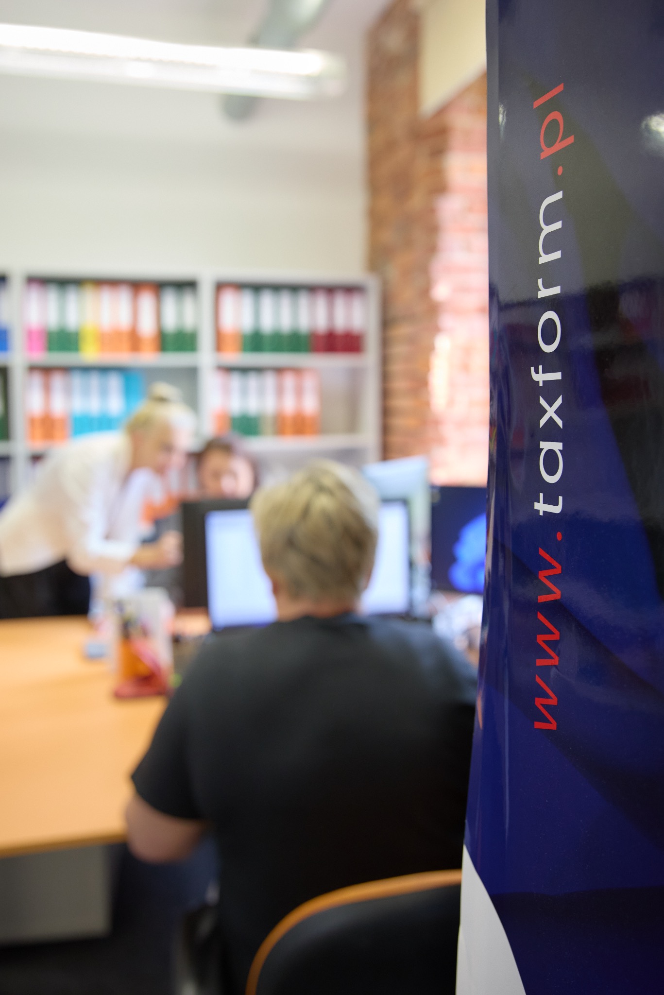 Biuro, w tle regał z segregatorami, pracownicy przy biurku, w pierwszym planie baner reklamowy z adresem strony internetowej.