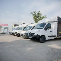 Szereg białych samochodów dostawczych ustawionych na żwirowym parkingu, z logo 'AGRO MIX' widocznym na jednym z nich, w tle budynki i drzewa pod błękitnym niebem.