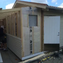 Budowa drewnianej dobudówki: montaż desek elewacyjnych na konstrukcji szkieletowej z izolacją, widoczne narzędzia i fragment istniejącego budynku z białą stolarką drzwiową.