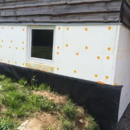 Ocieplenie ściany budynku styropianem z widocznymi kołkami mocującymi, czarna folia kubełkowa przy gruncie, drewniana elewacja górnej części ściany, trawa i ziemia przed budynkiem.