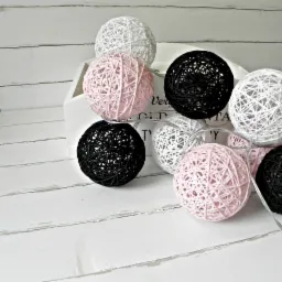 Dekoracyjne cotton balls w kolorach: białym, czarnym i różowym, ułożone na białej, drewnianej skrzynce i desce. Delikatne światło tworzy przytulny nastrój.