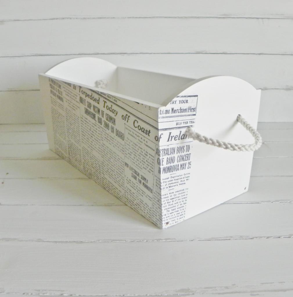 Biały pojemnik dekoracyjny z motywem gazetowym i uchwytami z grubego sznurka, stojący na białych deskach. Element wystroju wnętrza w stylu vintage.