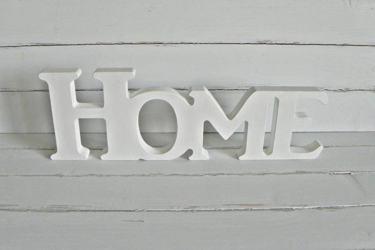 Biały, dekoracyjny napis 'HOME' na tle szarej, drewnianej ściany. Litery wykonane z drewna, o wyraźnej fakturze. Minimalistyczny design, stonowana kolorystyka.