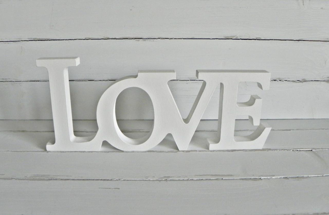 Biały napis 'LOVE' z drewna na tle białych, pomalowanych desek. Dekoracja wnętrza, subtelne światło, prosty design.