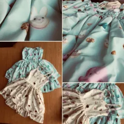 Dwie sukienki dziecięce z motywem słodyczy i zwierząt, jedna w pastelowym błękicie, druga w kremowym odcieniu, leżące na drewnianym blacie.