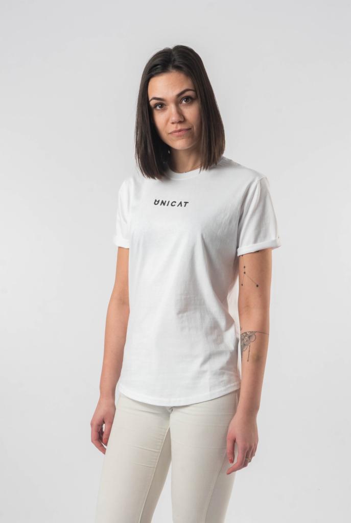Kobieta pozuje w białym T-shircie z czarnym napisem 'UNICAT' na tle jasnego studia, z widocznym tatuażem na ramieniu.