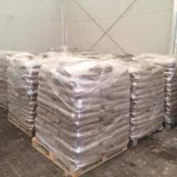 Spiętrzone, owinięte folią worki na paletach w magazynie. Widoczne drewniane palety i betonowa podłoga. Worki ułożone w rzędach pod białym stropem.