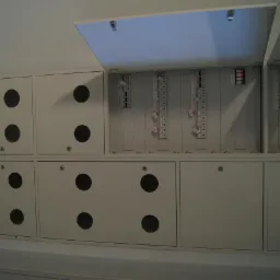 Szafka rozdzielcza z otwartymi drzwiczkami, widoczne listwy zaciskowe i elementy instalacji elektrycznej, dolne drzwiczki z okrągłymi otworami wentylacyjnymi.