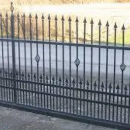 Czarna, metalowa brama wjazdowa z dekoracyjnymi elementami w kształcie rombów, zwieńczona ostrymi grotami, na tle asfaltowej drogi i zielonego pola.