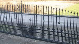 Czarna, metalowa brama wjazdowa z dekoracyjnymi elementami w kształcie rombów, zwieńczona ostrymi grotami, na tle asfaltowej drogi i zielonego pola.