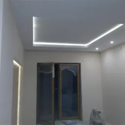 Nowoczesny sufit podwieszany z oświetleniem LED w kształcie prostokąta i punktowym, widoczne drzwi zabezpieczone folią i worek gipsu na podłodze.