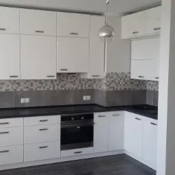 Biała kuchnia z połyskiem, ciemny blat i mozaika na ścianie. Nowoczesny design z oświetleniem punktowym. Widok na zabudowę kuchenną od frontu.