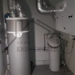 Szary pionowy zasobnik ciepłej wody użytkowej z niebieskim elementem u góry, podłączony do rur wentylacyjnych w pomieszczeniu technicznym z szarą podłogą i widocznym zmiękczaczem wody po prawej...