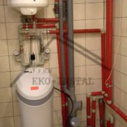 Biały zmiękczacz wody EKO-INSTAL obok czerwonych rur instalacji grzewczej w pomieszczeniu wyłożonym kwadratowymi płytkami.