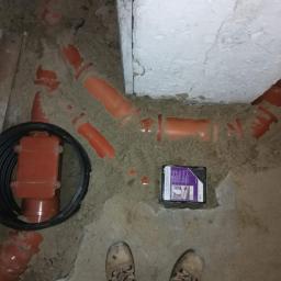 A&L INSTALATOR Leszek Szrajber - Wykop z ułożonymi rurami kanalizacyjnymi z tworzywa w kolorze pomarańczowym, czarna rura karbowana, pudełko z produktem i buty osoby wykonującej prace. Podłoże piaszczyste.