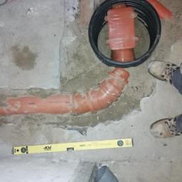 A&L INSTALATOR Leszek Szrajber - Pomarańczowa rura kanalizacyjna z kolankiem, żółta poziomica Deora POP oraz czarny właz rewizyjny w szarej betonowej posadzce, widoczne buty i nogawki pracownika.