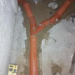 A&L INSTALATOR Leszek Szrajber - Pomarańczowe rury kanalizacyjne w kształcie litery Y, częściowo zasypane piaskiem, widoczne w wykopie, obok poziomnica i but robotnika.