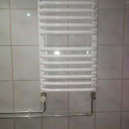 Biały, drabinkowy grzejnik łazienkowy zamontowany na ścianie z kwadratowych, jasnych płytek, z widocznymi rurami doprowadzającymi wodę i zaworem termostatycznym.