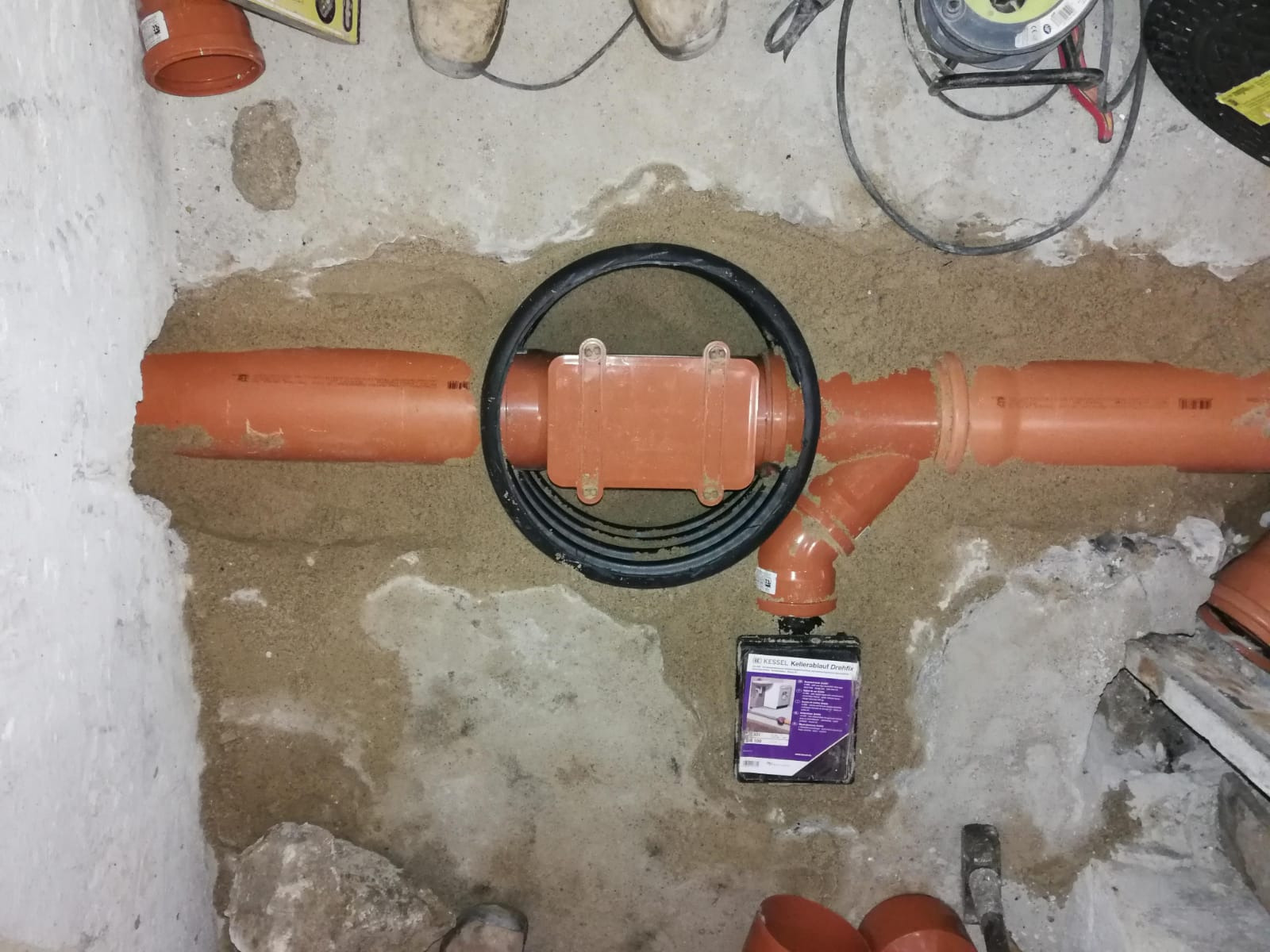 Pomarańczowe rury kanalizacyjne ułożone w piasku, z czarną uszczelką i pudełkiem produktu Kessel Kellerablauf Drehfix, widoczne elementy instalacji podłogowej.