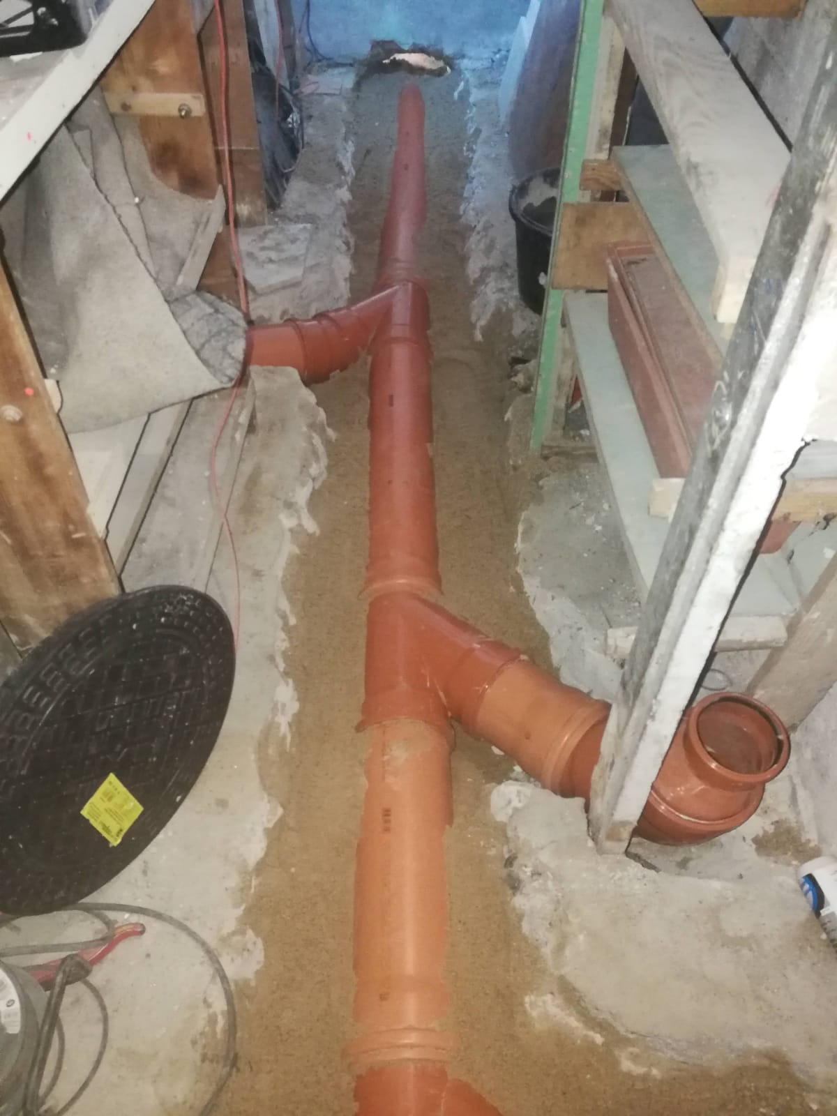 Pomarańczowe rury kanalizacyjne ułożone w wykopie w piasku, widoczna studzienka rewizyjna z żółtą naklejką i fragment drabiny.