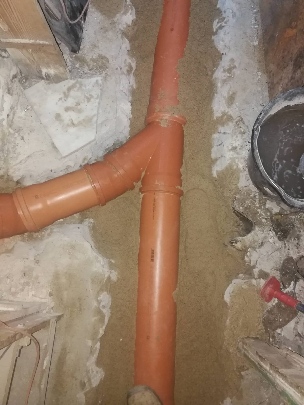 Pomarańczowe rury kanalizacyjne ułożone w wykopie zasypanym piaskiem, widoczny fragment ściany z resztkami zaprawy i wiadro z zaprawą obok.