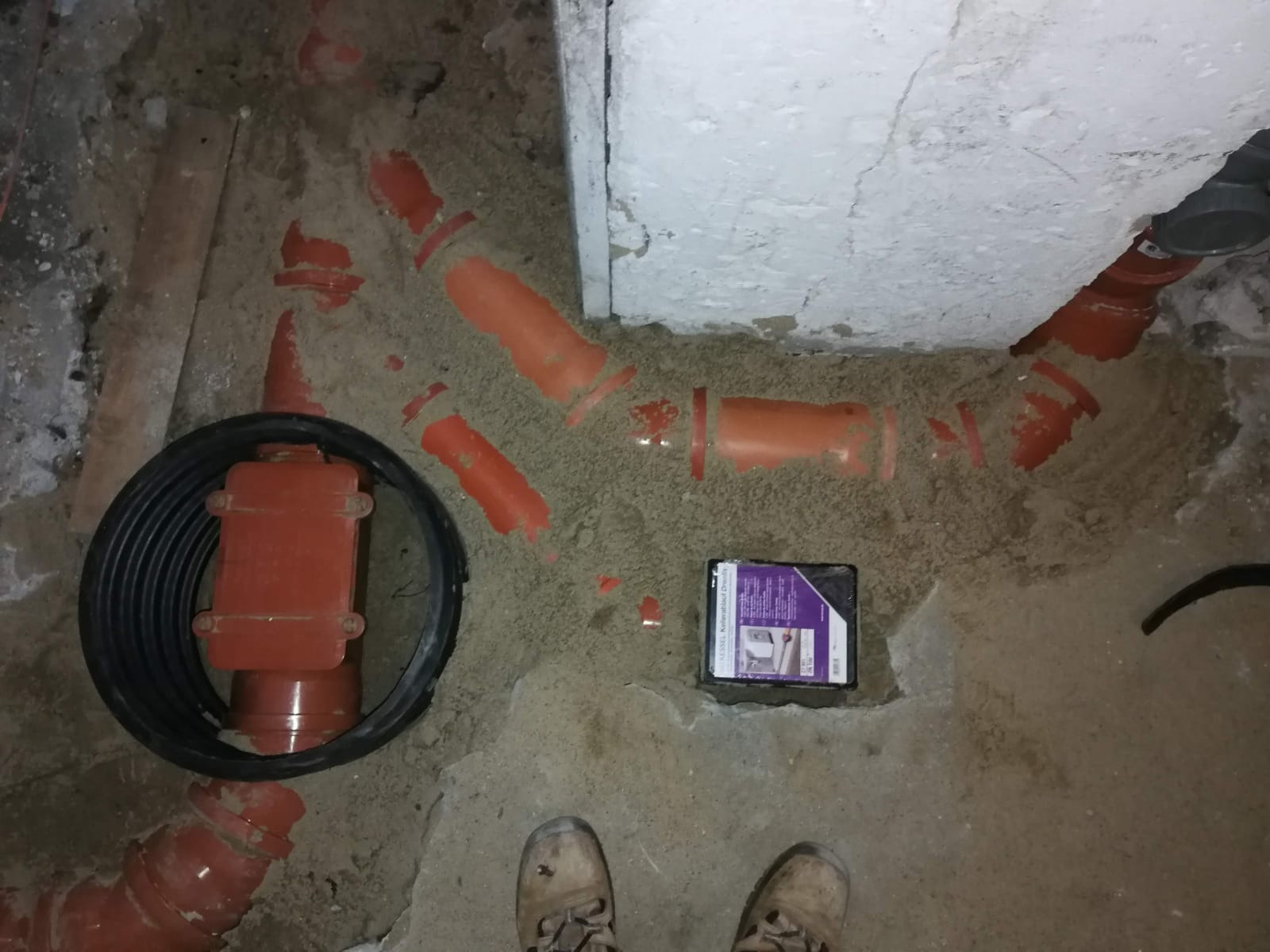 Wykop z ułożonymi rurami kanalizacyjnymi z tworzywa w kolorze pomarańczowym, czarna rura karbowana, pudełko z produktem i buty osoby wykonującej prace. Podłoże piaszczyste.