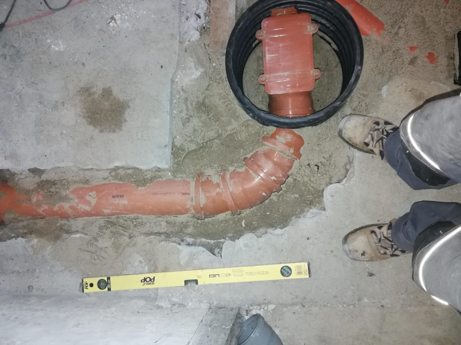 Pomarańczowa rura kanalizacyjna z kolankiem, żółta poziomica Deora POP oraz czarny właz rewizyjny w szarej betonowej posadzce, widoczne buty i nogawki pracownika.