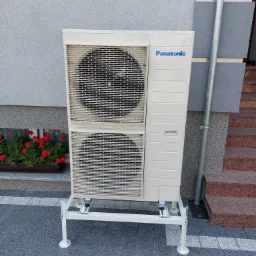 Zewnętrzna jednostka klimatyzacyjna Panasonic na białym stelażu, ustawiona przy szarej ścianie budynku z czerwonymi kwiatami w doniczce w tle i granitowymi schodami z prawej strony.