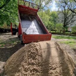 Czerwona wywrotka z otwartą skrzynią ładunkową, z której wysypano dużą pryzmę piasku na trawnik w otoczeniu drzew.