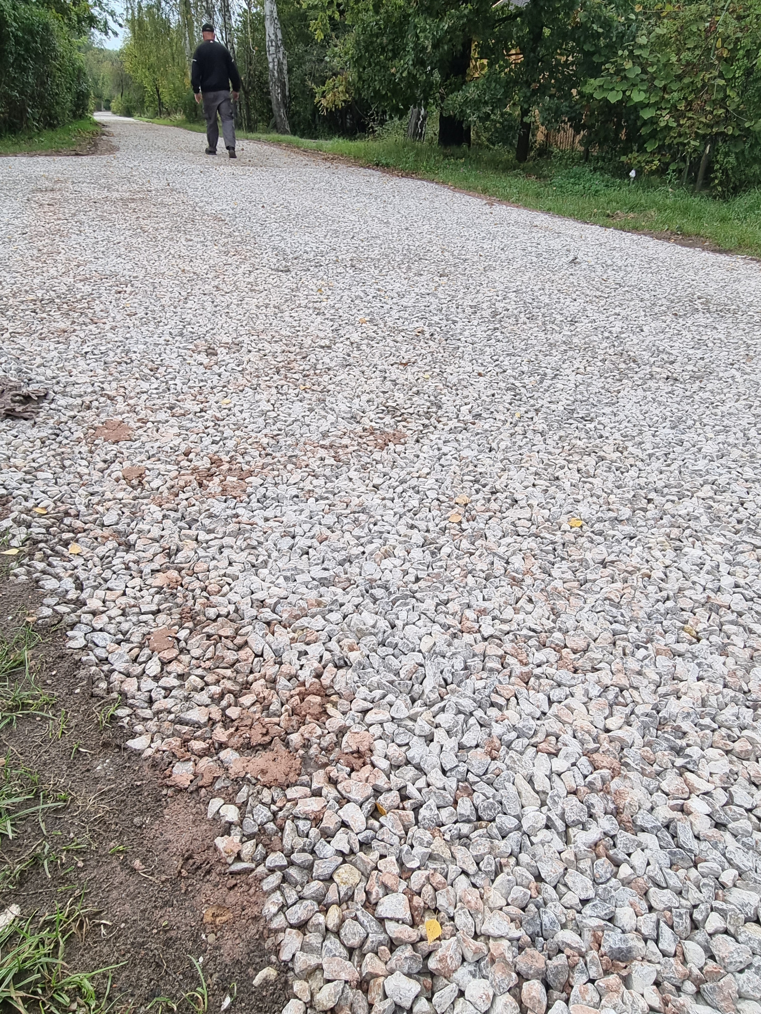 Utwardzona ścieżka żwirowa w parku, z widocznym fragmentem ziemi po bokach i osobą spacerującą w oddali.