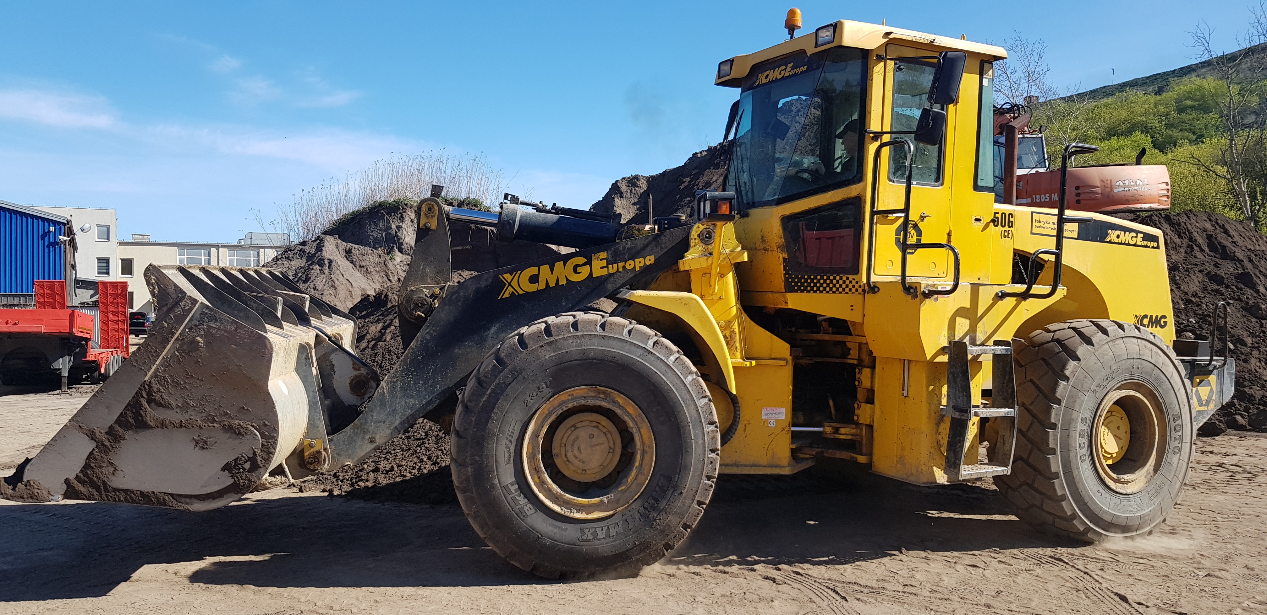 Żółta ładowarka kołowa XCMG 50G z łyżką załadowaną ziemią na placu budowy w słoneczny dzień, w tle budynki i hałda ziemi.