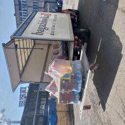 YELLOW TRANSPORT ROBERT ŻÓŁTY - Otwarta paka ciężarówki z zabawkami w folii, gotowa do transportu. Widoczny budynek w tle i komin z dymem. Słoneczny dzień.