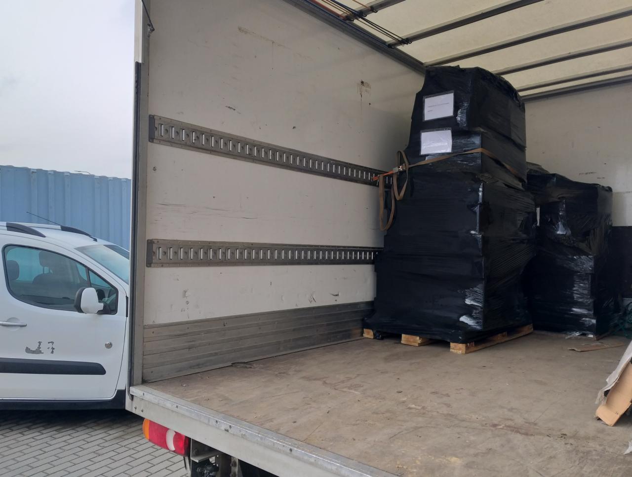 Wnętrze otwartej naczepy z ładunkiem zabezpieczonym czarną folią stretch na paletach, widoczne szyny mocujące i częściowo biały samochód dostawczy na zewnątrz.