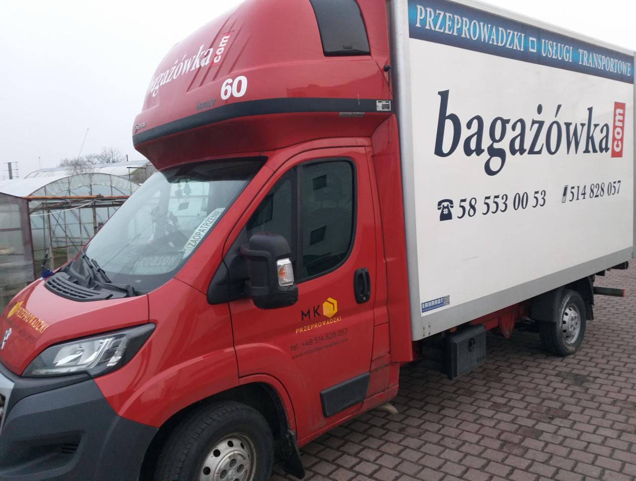 Czerwony samochód dostawczy z białą zabudową, oznaczony logo 'bagażówka.com' i numerami telefonów, zaparkowany na kostce brukowej, z widocznymi szklarniami w tle.