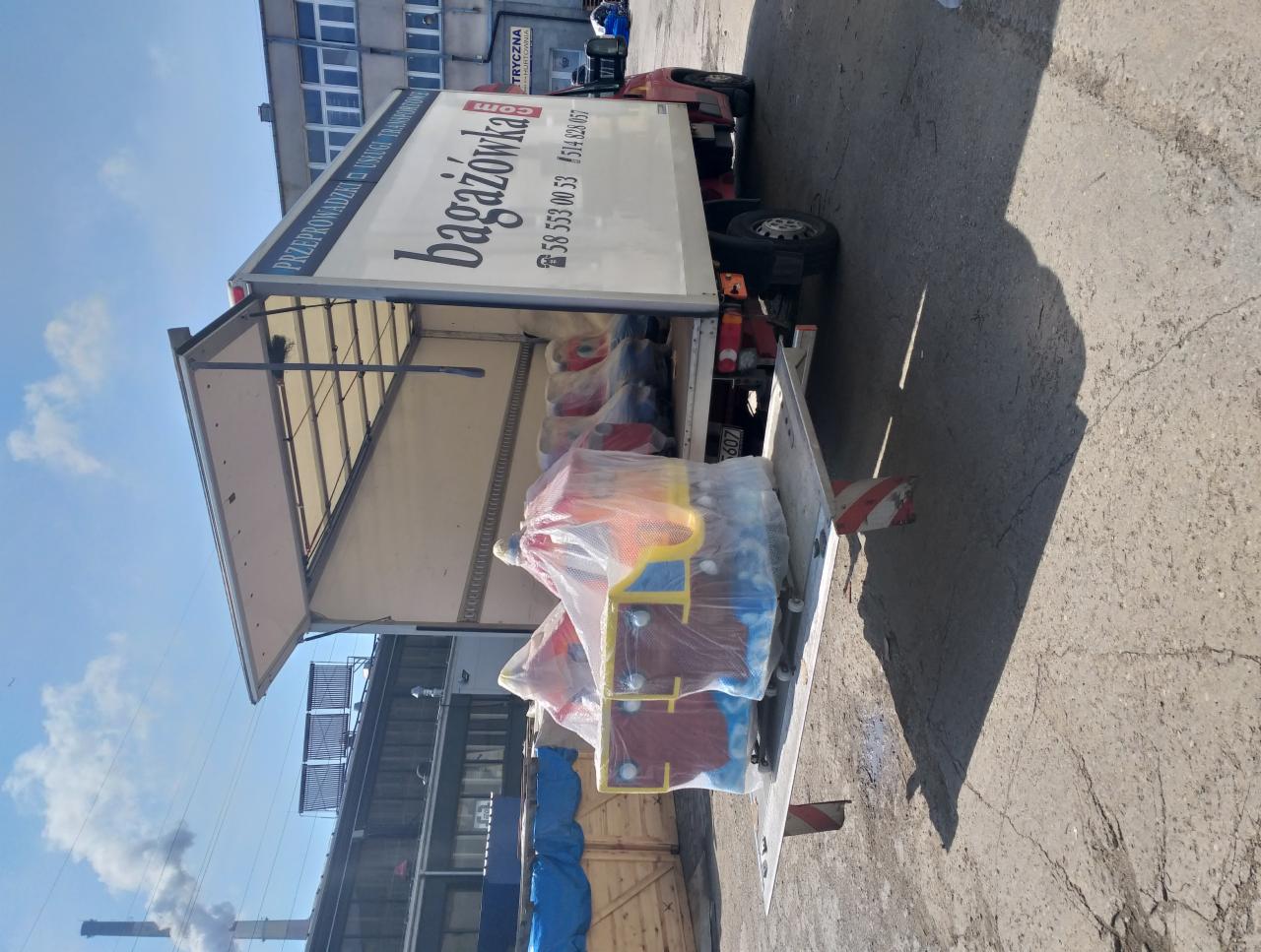 Otwarta paka ciężarówki z zabawkami w folii, gotowa do transportu. Widoczny budynek w tle i komin z dymem. Słoneczny dzień.