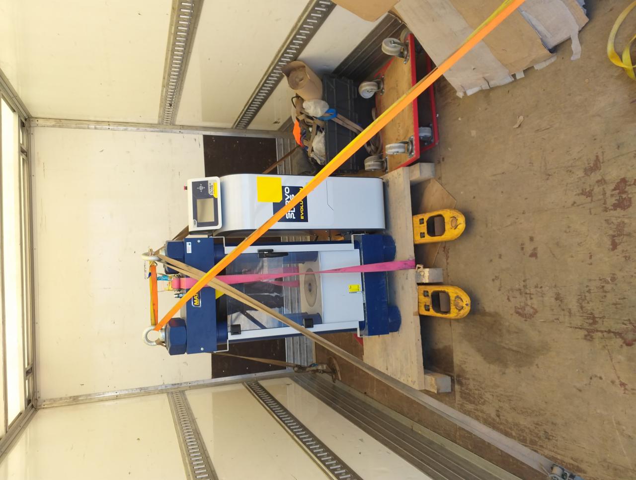 Maszyna na palecie zabezpieczona pasami transportowymi wewnątrz pojazdu. Widoczne wózki transportowe i kartony. Transport specjalistyczny.