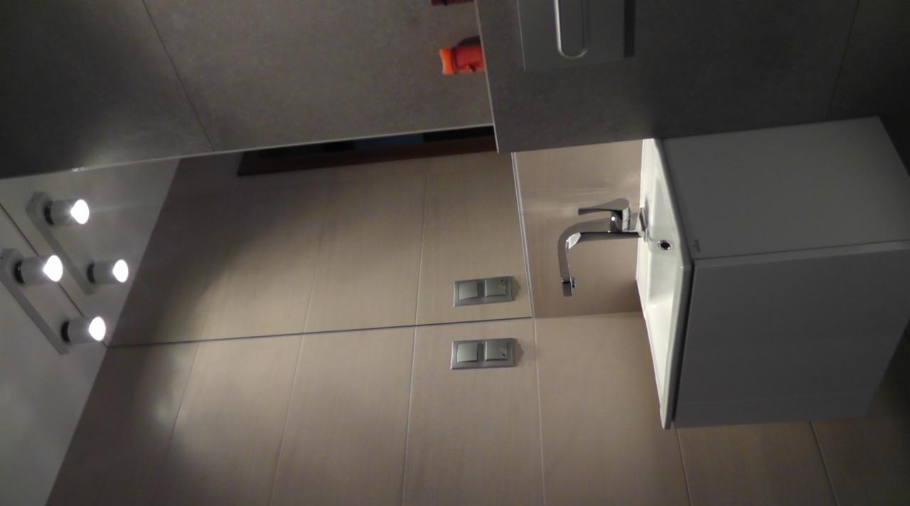 Wykończona łazienka w beżowych płytkach z umywalką, baterią i lustrem z oświetleniem. Widoczne dwa włączniki światła. Ujęcie z dołu na górę, perspektywa z narożnika.